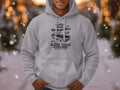 A person stands outdoors wearing a Garment Graphics classic fit gray hoodie featuring a London city vintage car and classic car club design. Snow falls softly, with blurred lights and trees creating a picturesque scene ideal for automobile enthusiasts.