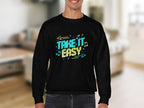 A person is wearing a Garment Graphics black sweatshirt with vibrant Take It Easy text and stylish graphics. The background shows a softly blurred interior with a cozy sofa and wall decor, while unisex sizing ensures comfort for all.