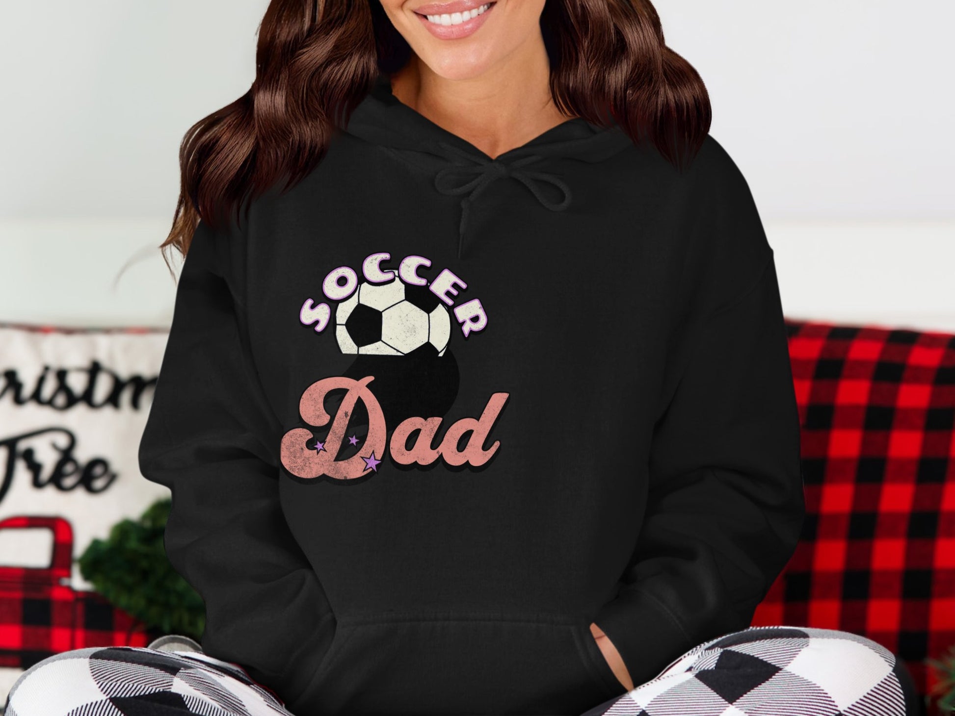 A person, partly shown, is seated casually wearing a black Soccer Dad hoodie with a soccer ball graphic from Garment Graphics. The scene is cozy with a red and black checkered pillow in the background.