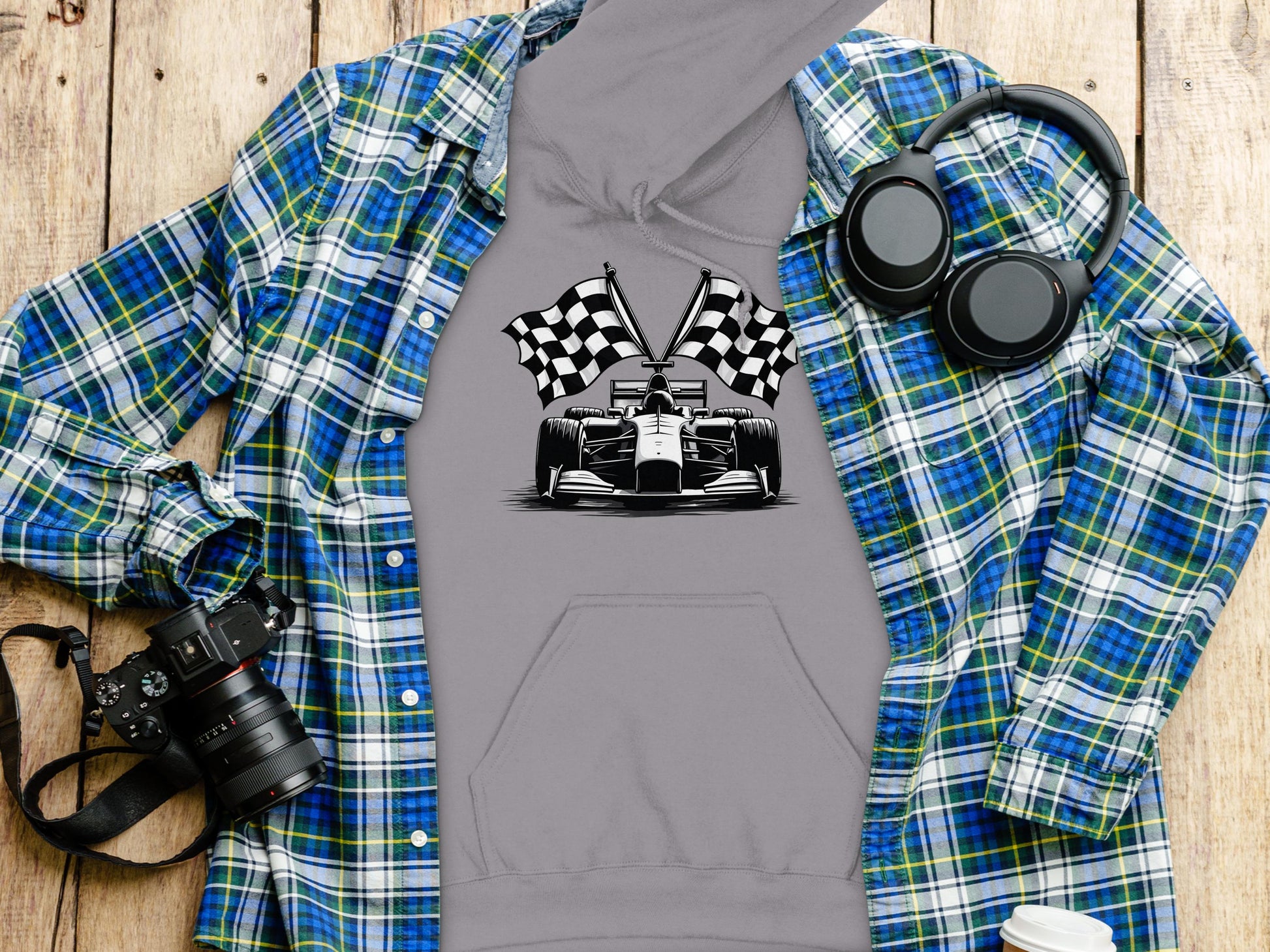 A Garment Graphics gray hoodie with a race car and checkered flag design is on a blue and green plaid shirt. Next to it are racing-themed headphones, a camera, and a white cup on wood.