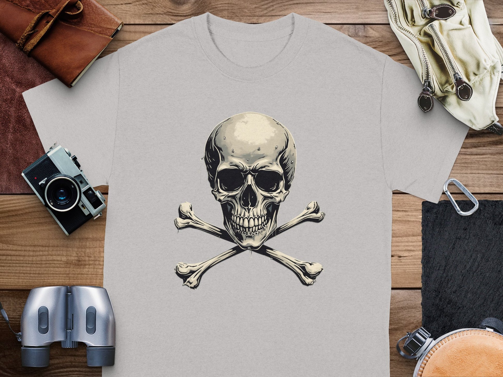 On a wooden surface, Garment Graphics graphic t-shirt with a striking skull and crossbones design is paired with a leather wallet, binoculars, vintage camera, and beige gloves for an intriguing fashion ensemble.