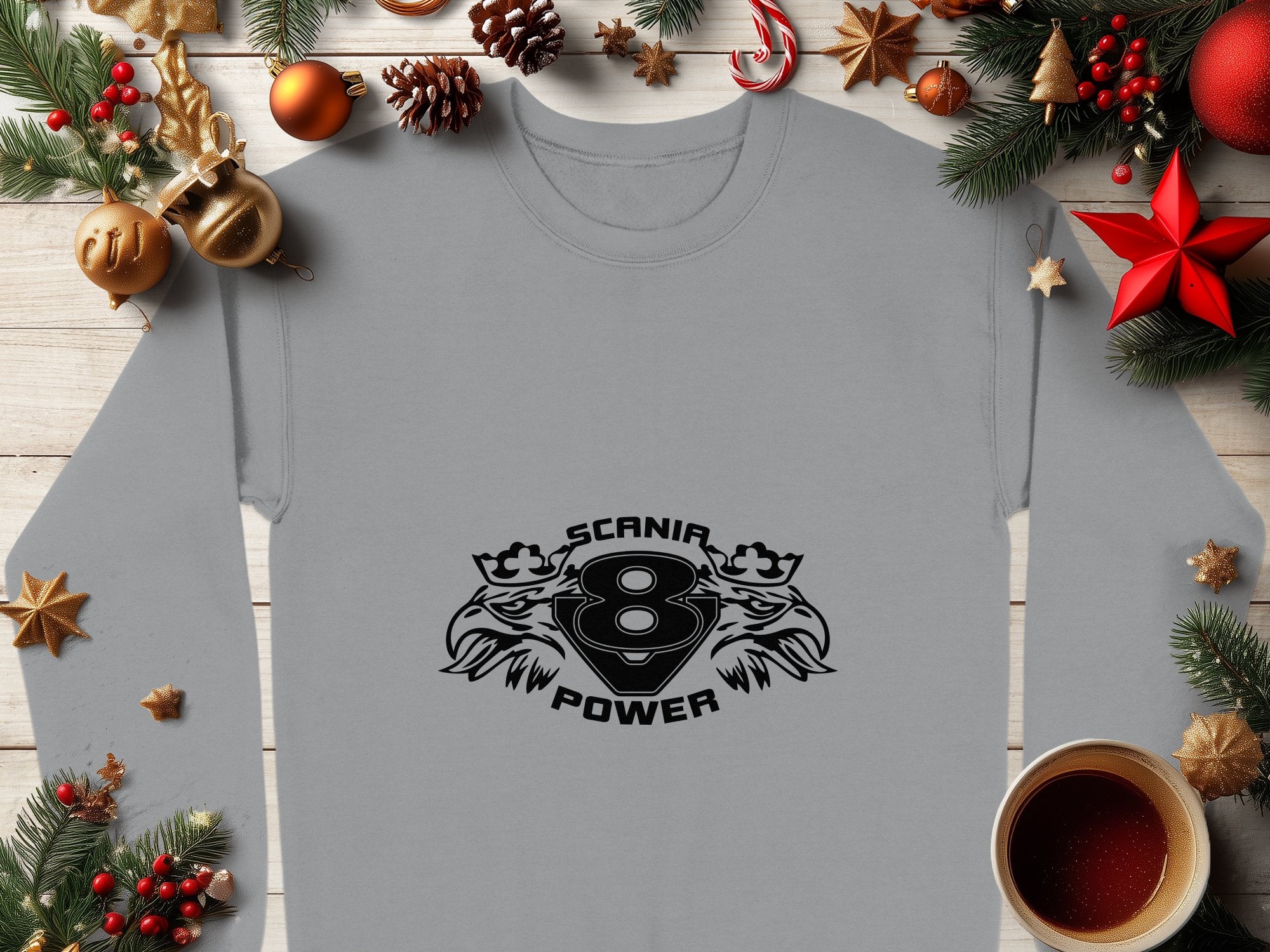 A Garment Graphics gray sweatshirt featuring the Scania Power 8 logo with crest-like animal heads is surrounded by festive Christmas decor—pine branches, ornaments, and a steaming coffee cup on wood—making it ideal cozy attire for the season.