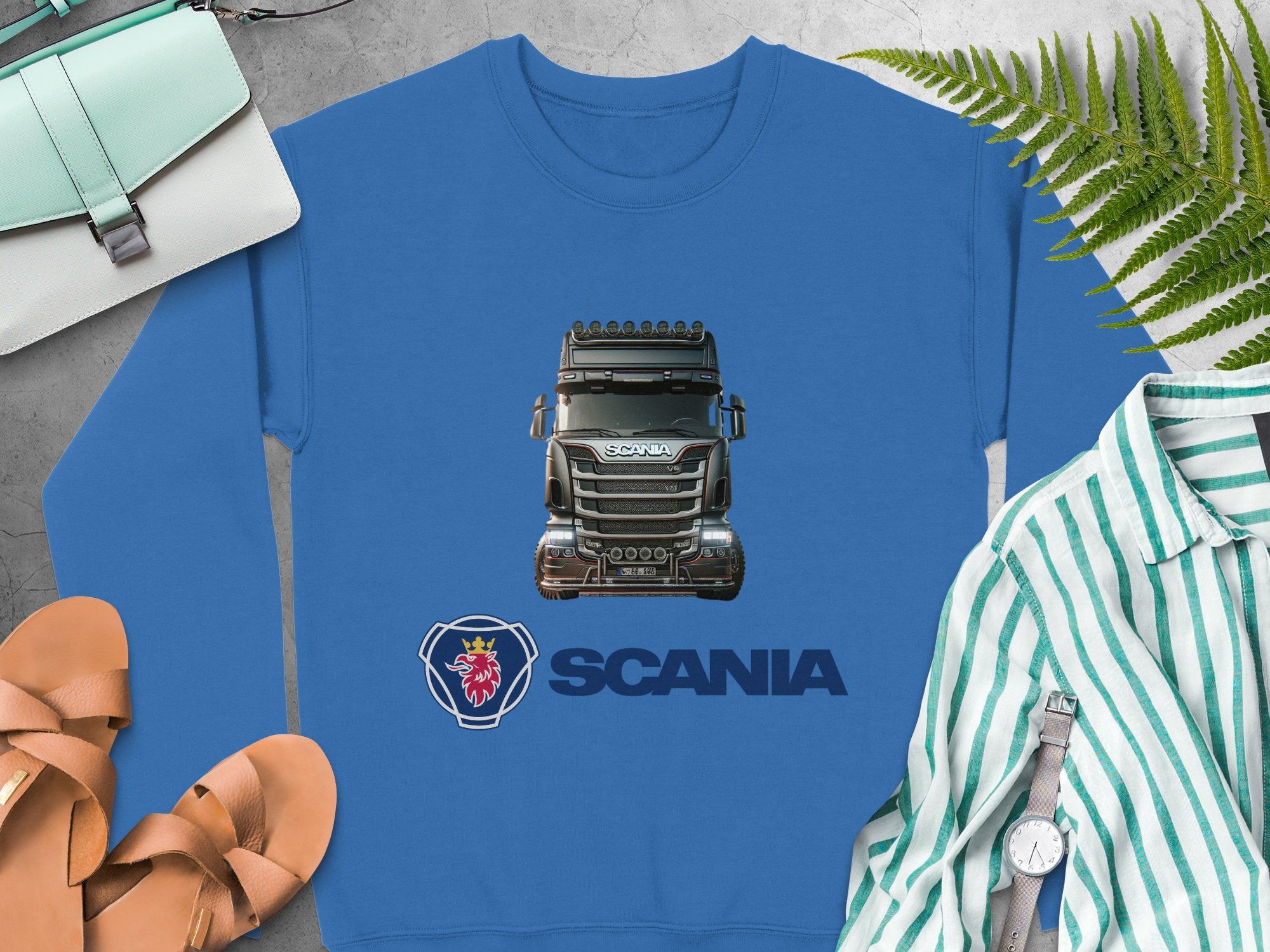Garment Graphics blue graphic crewneck sweatshirt features a Scania truck and logo. Surrounding it are plants, sandals, a watch, a striped shirt, and a small handbag on gray—the perfect piece for truck enthusiasts.
