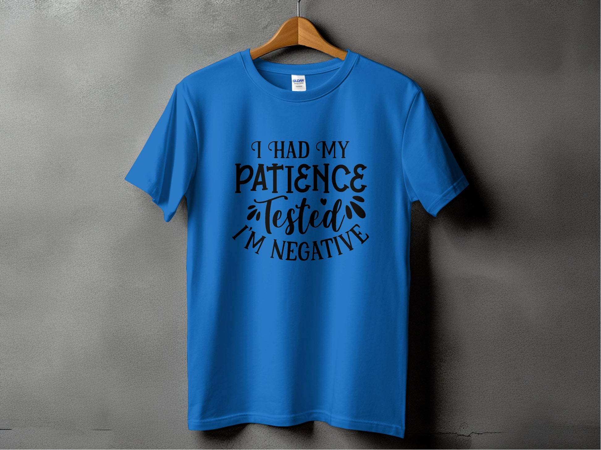 A blue t-shirt from Garment Graphics with a sarcastic attitude hangs on a wooden hanger against a gray wall. It humorously states, I Had My Patience Tested Im Negative in black text with decorative flourishes.