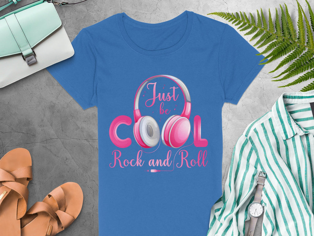 Garment Graphics blue t-shirt showcases headphones with the bold Just be Cool Rock and Roll text, alongside a mint green purse, brown sandals, and a striped shirt complimented by a watch. A fern completes the ensemble on a gray textured backdrop. Ideal for music lovers!.