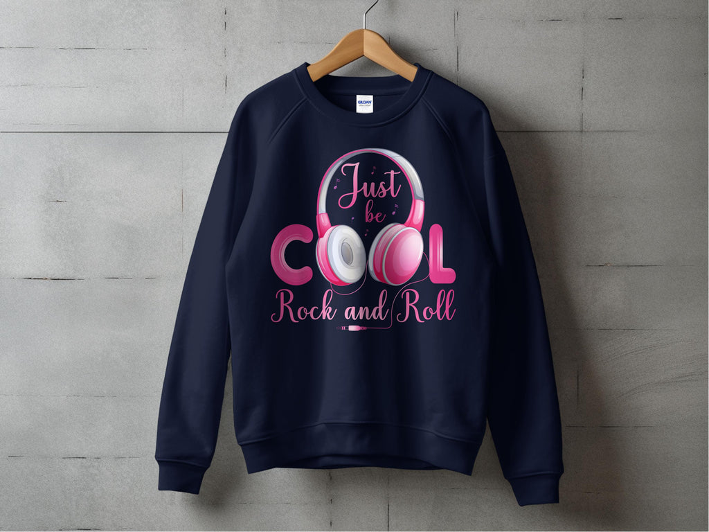 A navy Garment Graphics sweatshirt with the Just be COOL Rock and Roll pink and white headphones design hangs on a wooden hanger. Its a chic statement for music lovers, set against a gray concrete backdrop to highlight the Rock and Roll motif.