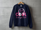 A navy Garment Graphics sweatshirt with the Just be COOL Rock and Roll pink and white headphones design hangs on a wooden hanger. Its a chic statement for music lovers, set against a gray concrete backdrop to highlight the Rock and Roll motif.