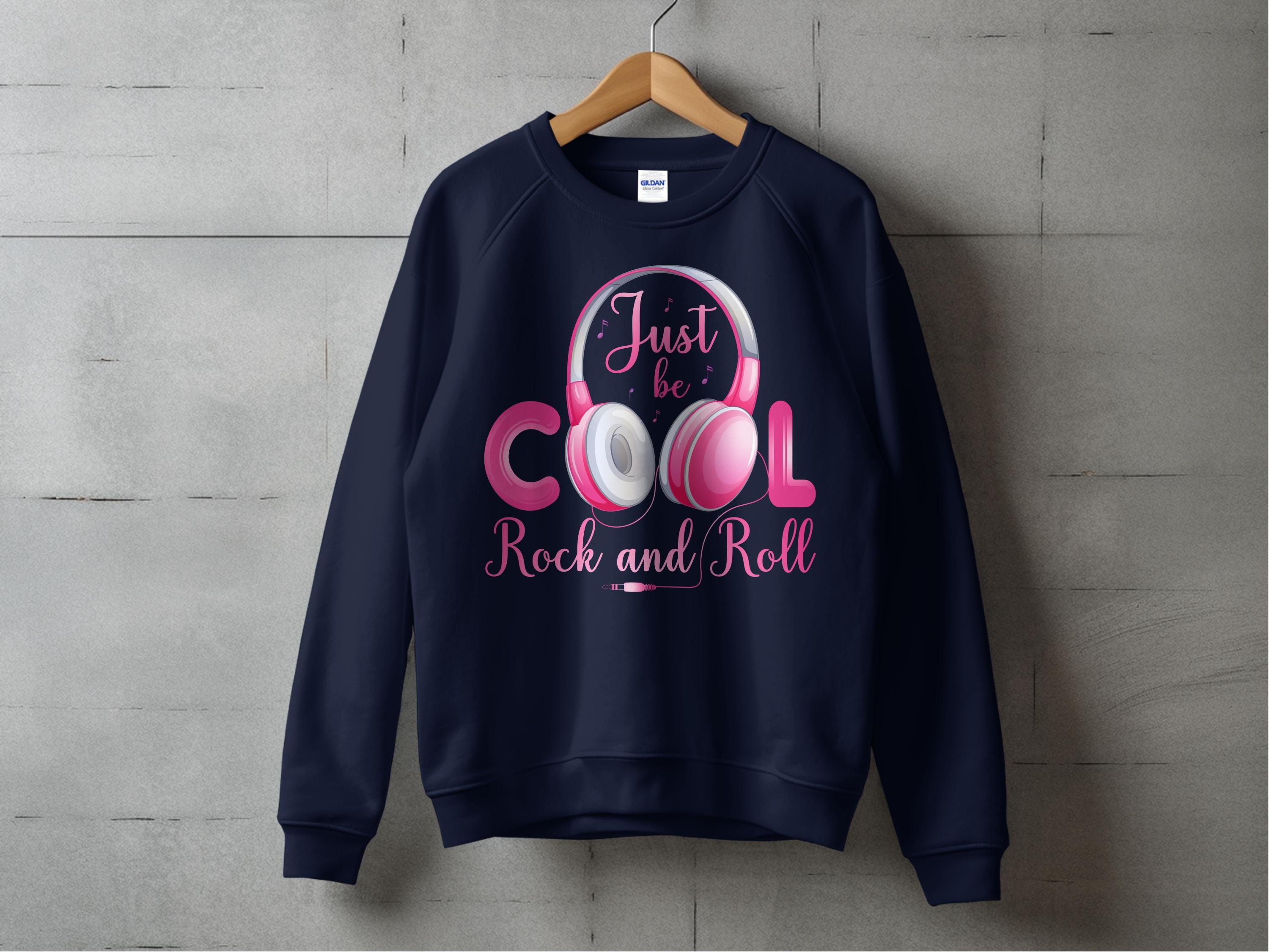 A navy Garment Graphics sweatshirt with the Just be COOL Rock and Roll pink and white headphones design hangs on a wooden hanger. Its a chic statement for music lovers, set against a gray concrete backdrop to highlight the Rock and Roll motif.