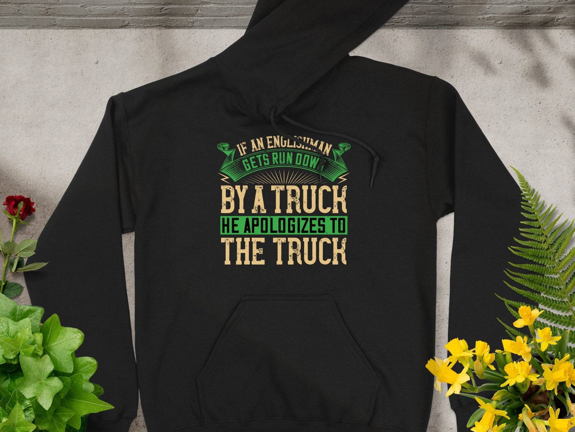 A black hoodie by Garment Graphics features a funny quote: If an Englishman gets run down by a truck, he apologizes to the truck in vibrant yellow, green, and beige lettering. Its displayed on concrete with red and yellow flowers and lush green leaves around it.