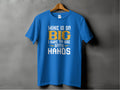 A classic fit blue tee from Garment Graphics, featuring bold white and yellow text reading, MINE IS SO BIG I HAVE TO USE BOTH HANDS, hangs on a wooden hanger against a gray background.