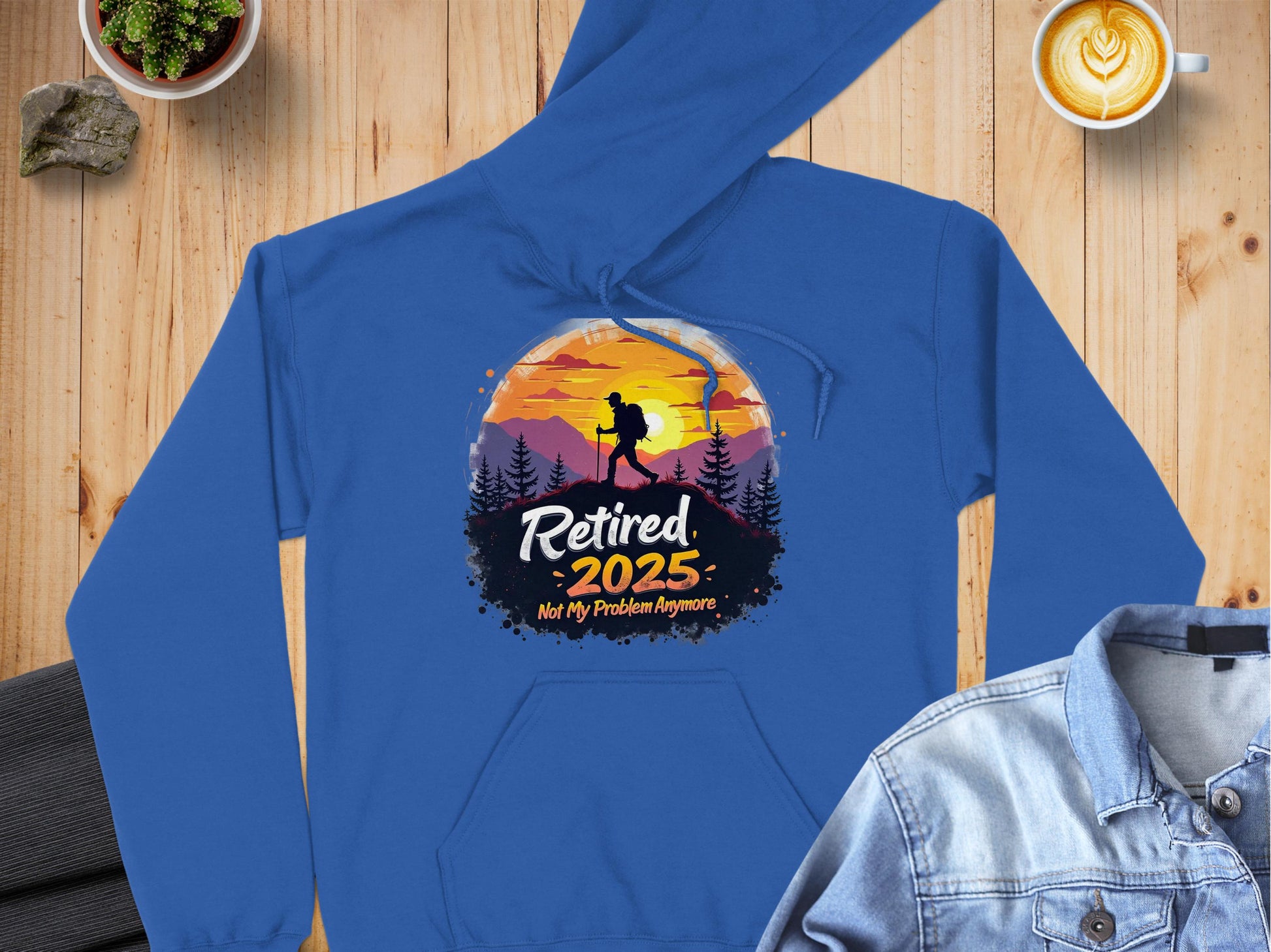 A Garment Graphics blue hoodie lies on a wooden surface, featuring a sunset mountain scene with a hiker, trees, and Retired 2025: Not My Problem Anymore. Nearby are a latte in a white cup, denim jacket, and potted plant. This hoodie embodies adventure and relaxation.