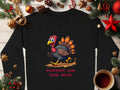 Garment Graphics black unisex shirt showcases a funny cartoon turkey running with Turkey on the Run printed below. It includes Christmas decorations like pine branches, ornaments, and a cozy cup of coffee on a wooden surface, adding festive fun to any wardrobe.