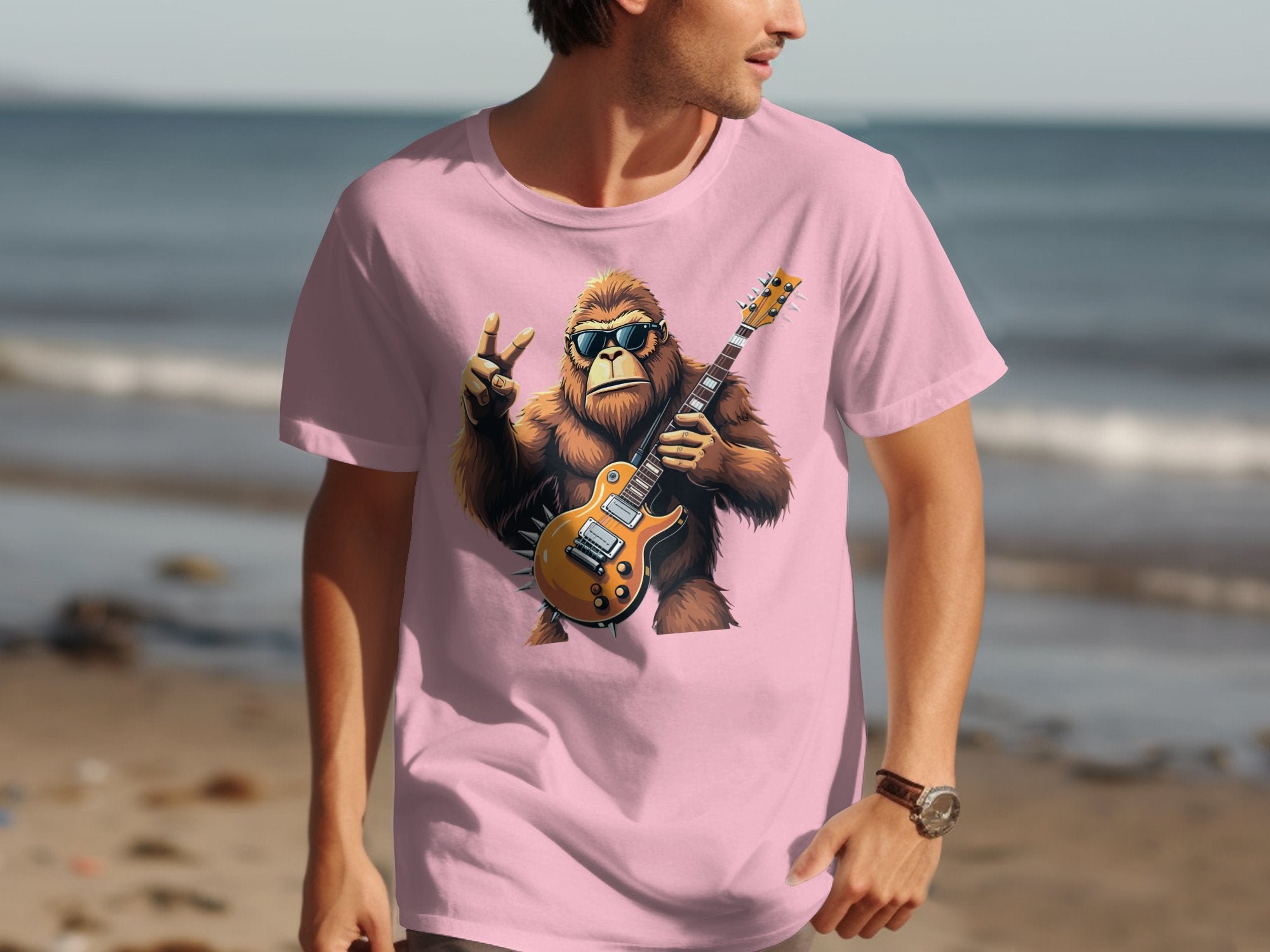 A man in a pink Garment Graphics t-shirt featuring a gorilla playing an electric guitar and making a peace sign strolls along the beach, with ocean waves creating the ideal setting for music enthusiasts.
