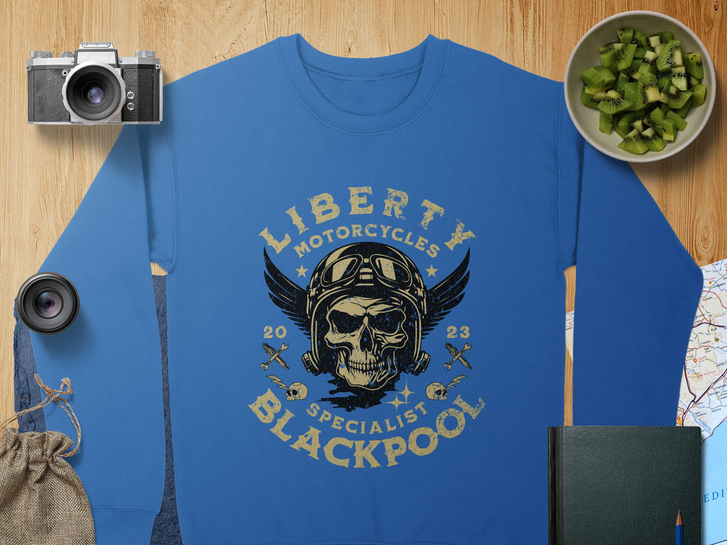 A blue sweatshirt by Garment Graphics showcases a skull in goggles with Liberty Motorcycles Specialist Blackpool 2023. The biker vibe is accentuated by a camera, lens, bag, book, map, and bowl of cucumber on a wooden surface.