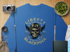 A blue sweatshirt by Garment Graphics showcases a skull in goggles with Liberty Motorcycles Specialist Blackpool 2023. The biker vibe is accentuated by a camera, lens, bag, book, map, and bowl of cucumber on a wooden surface.