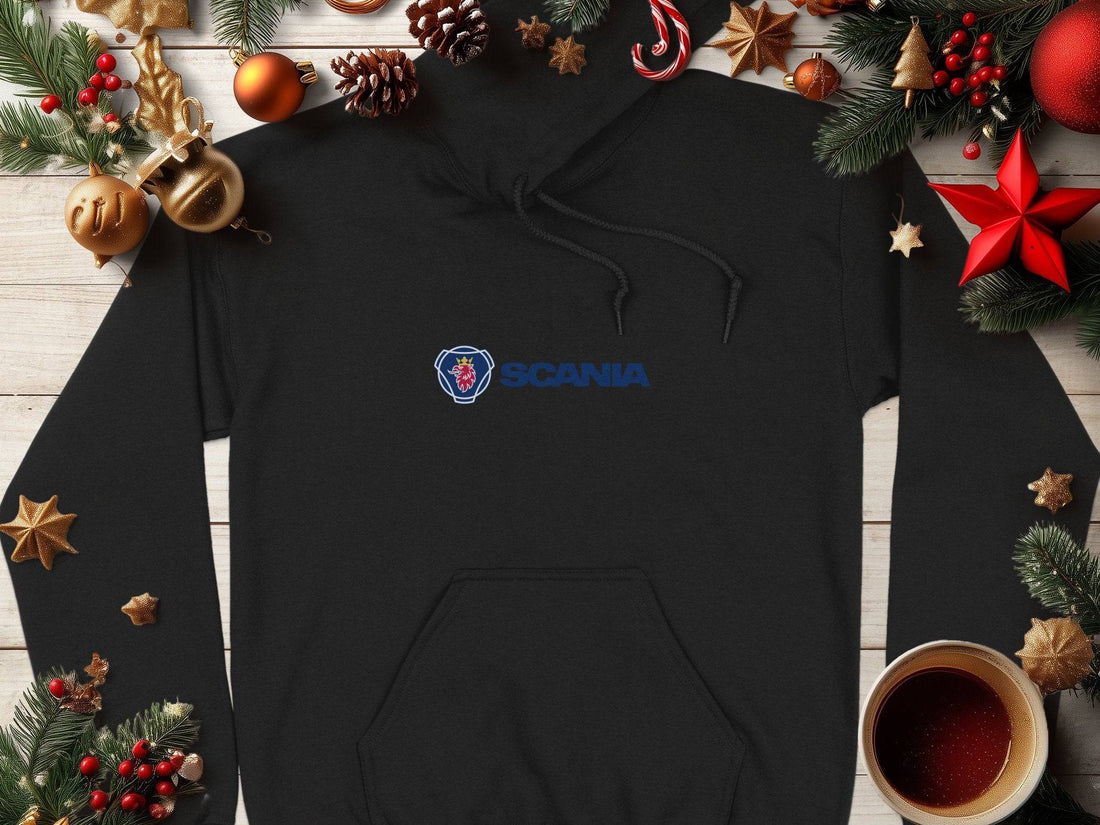 A black Garment Graphics hoodie with a blue Scania logo is displayed on wood, surrounded by festive pine cones, ornaments, stars, and a coffee cup—an exceptional fashion piece ideal for the seasons celebrations.