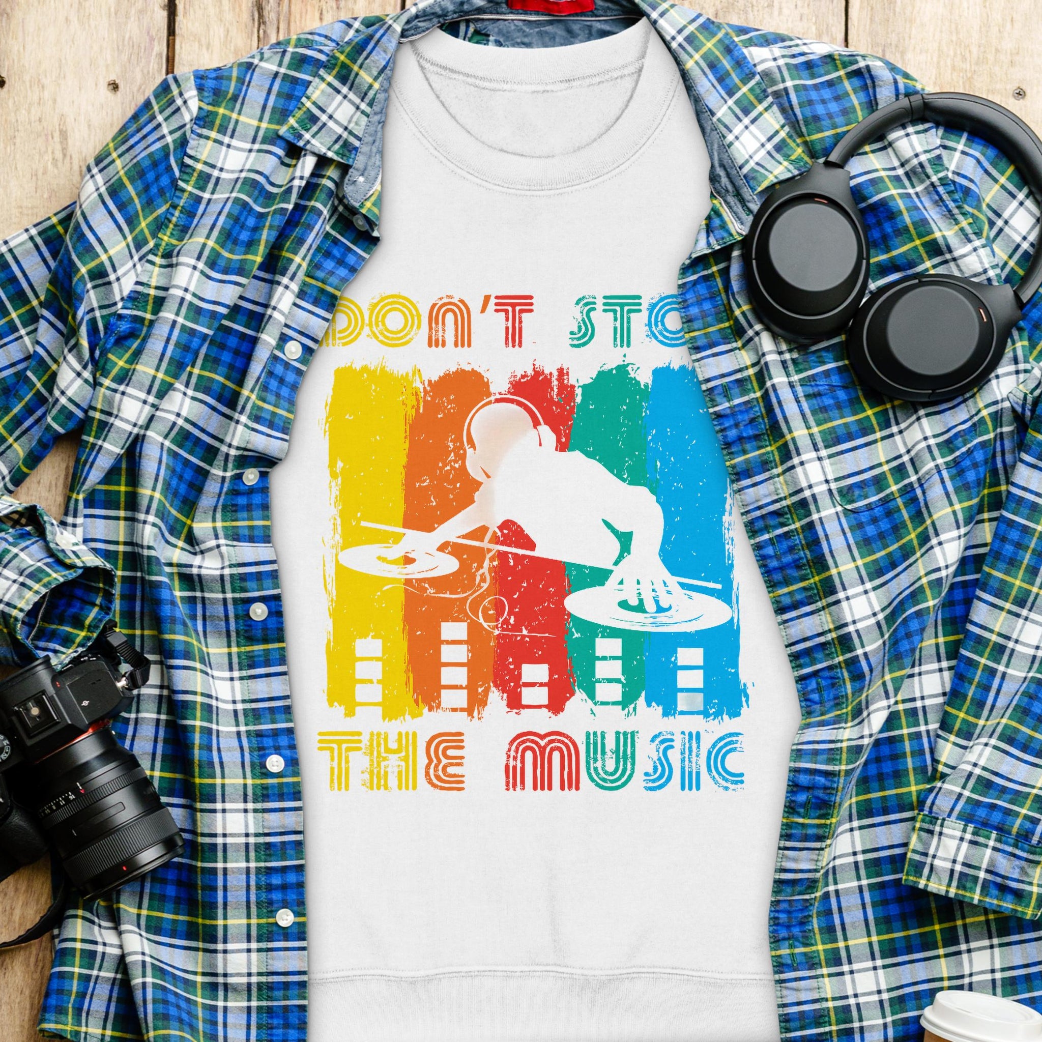 A DJ-themed t-shirt from Garment Graphics, featuring Dont Stop The Music, rests on a wooden surface. Its layered over a blue and green plaid shirt with black headphones, a digital camera, and a white cup with lid nearby. Available in unisex sizes for music lovers!.