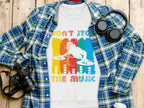 A DJ-themed t-shirt from Garment Graphics, featuring Dont Stop The Music, rests on a wooden surface. Its layered over a blue and green plaid shirt with black headphones, a digital camera, and a white cup with lid nearby. Available in unisex sizes for music lovers!.