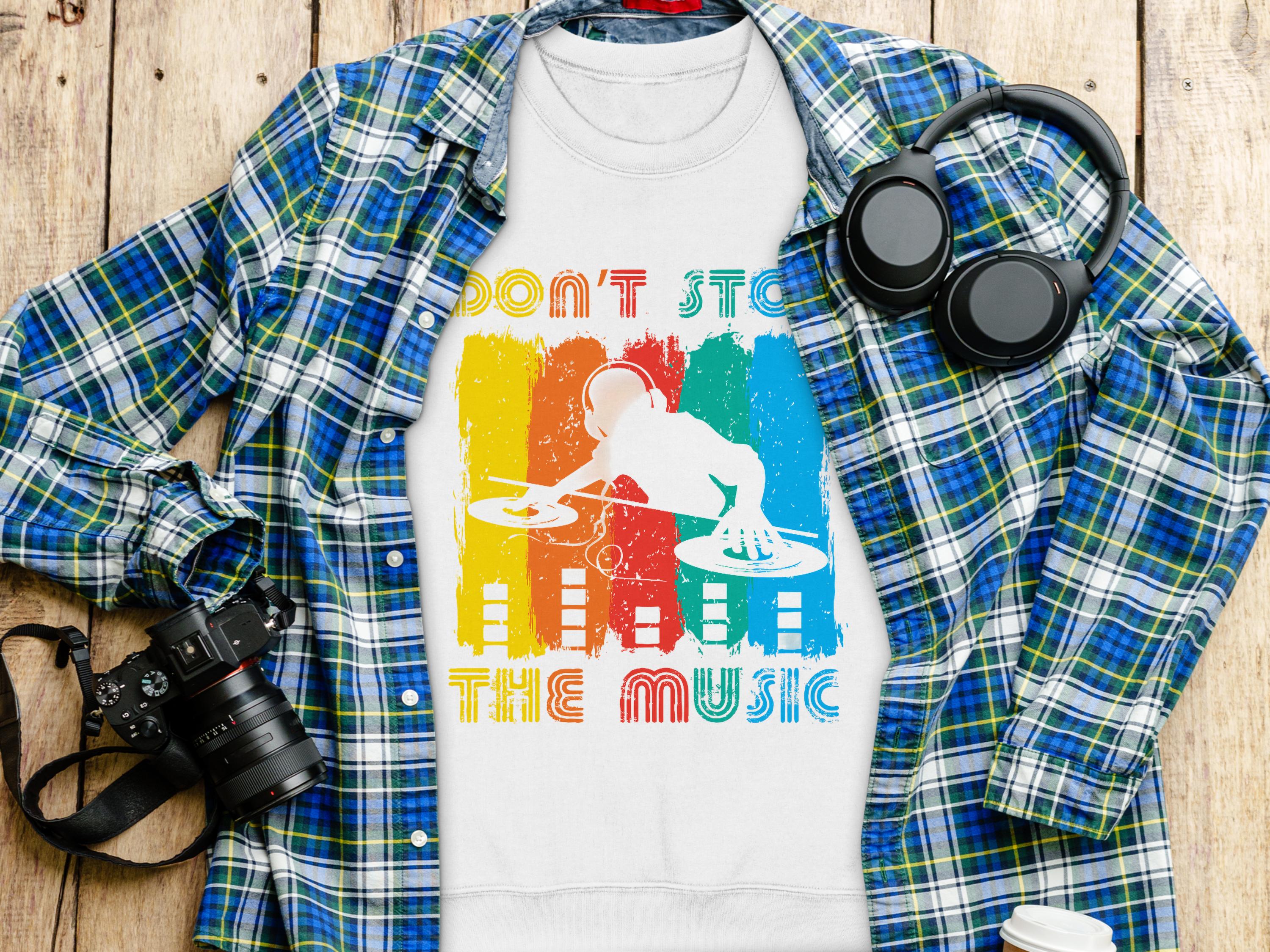 A DJ-themed t-shirt from Garment Graphics, featuring Dont Stop The Music, rests on a wooden surface. Its layered over a blue and green plaid shirt with black headphones, a digital camera, and a white cup with lid nearby. Available in unisex sizes for music lovers!.
