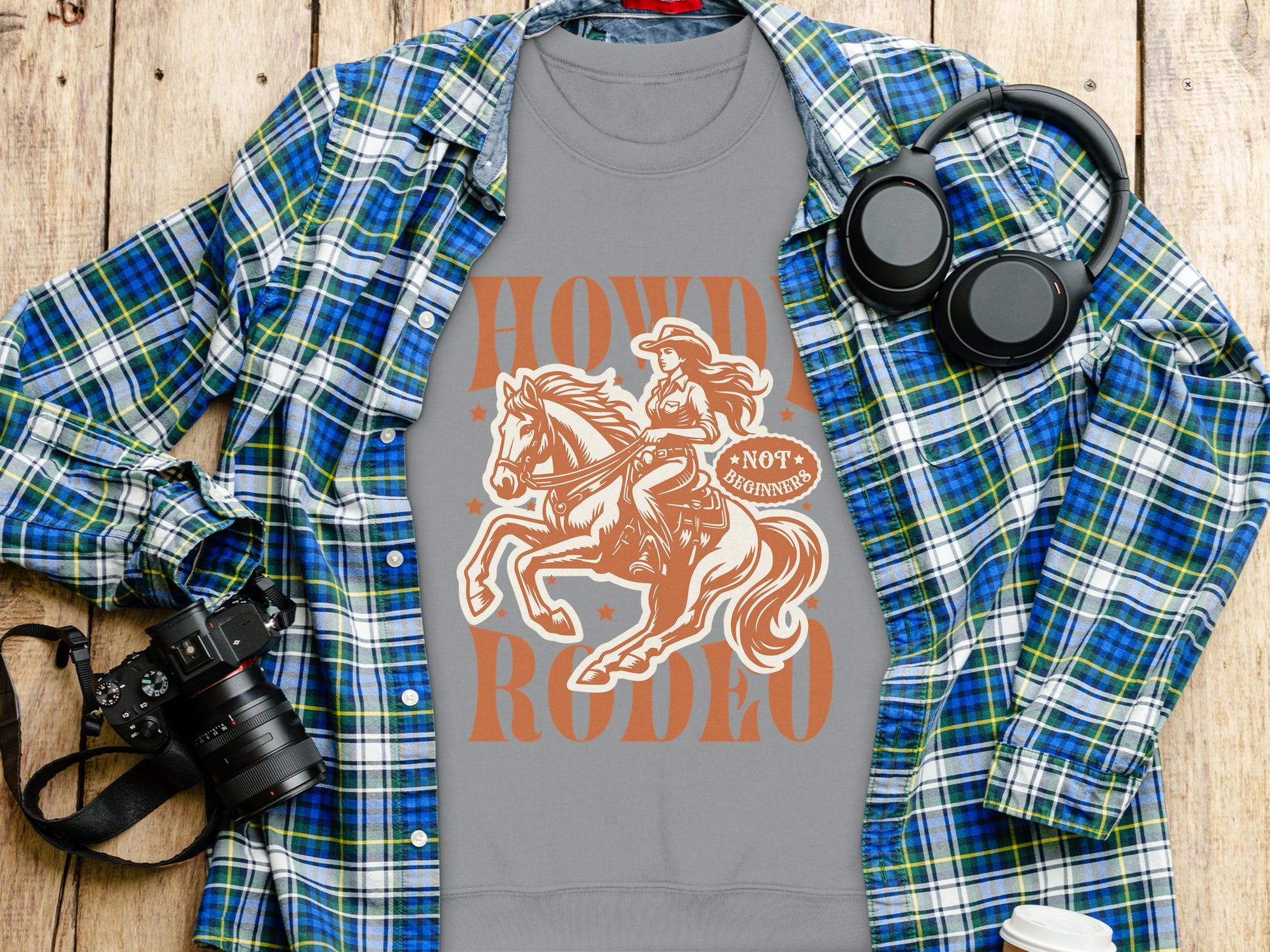 A Garment Graphics gray Howdy Rodeo cowboy and horse t-shirt lies beneath a blue and green plaid shirt, ideal for rodeo fans. Nearby, a camera, headphones, and a white cup sit on the wooden surface, embodying the cowboy spirit.