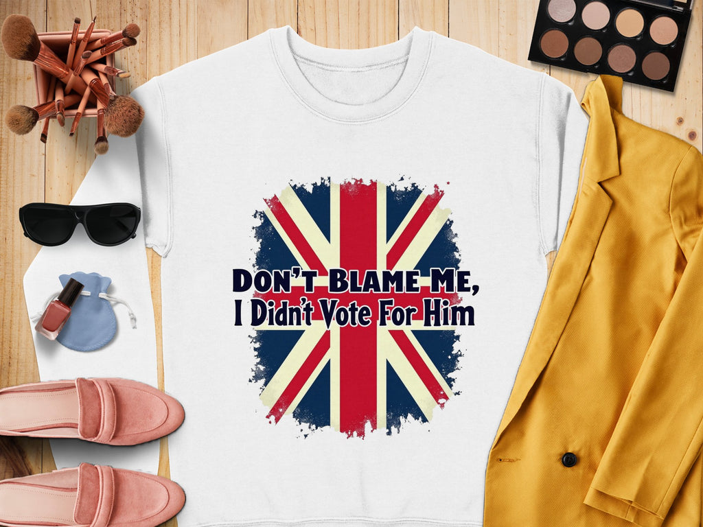This white T-shirt by Garment Graphics features a distressed Union Jack and the slogan, Dont Blame Me I Didnt Vote For Him. Styled with a yellow blazer, makeup brushes, sunglasses, pink loafers, lipstick, eyeshadow palette, and pouch. Unisex sizing ensures easy wearability.