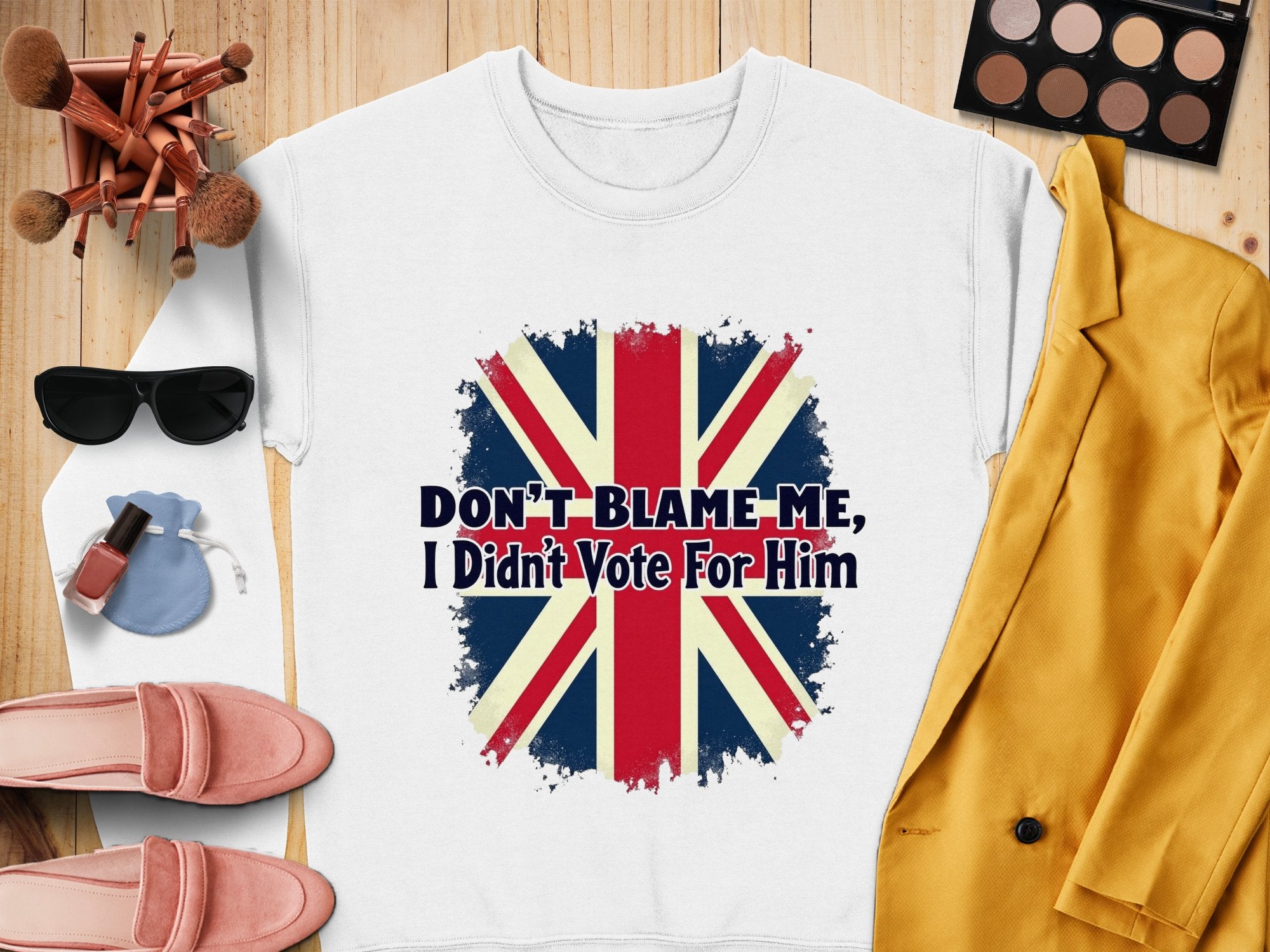 This white T-shirt by Garment Graphics features a distressed Union Jack and the slogan, Dont Blame Me I Didnt Vote For Him. Styled with a yellow blazer, makeup brushes, sunglasses, pink loafers, lipstick, eyeshadow palette, and pouch. Unisex sizing ensures easy wearability.