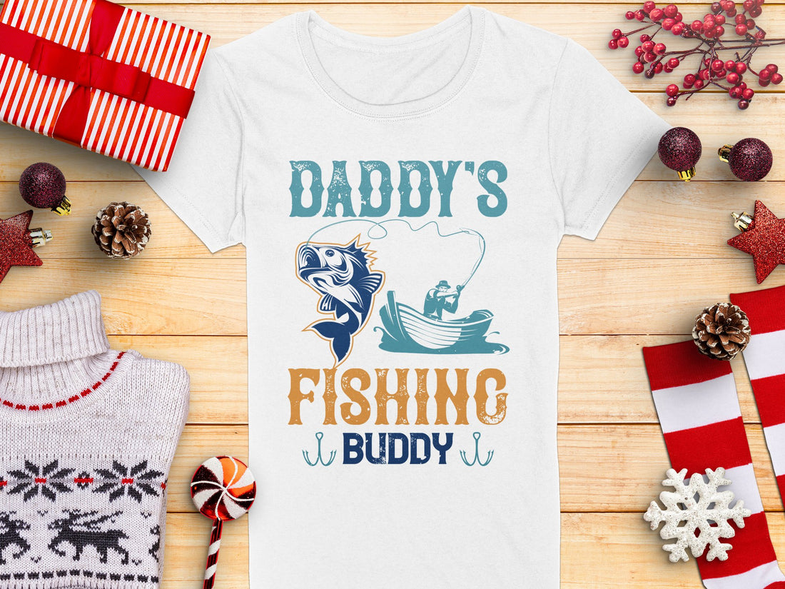 A Garment Graphics womens fishing shirt reads Daddys Fishing Buddy with a boat and fish illustration. Its displayed on a wooden surface with holiday decorations like wrapped gifts, pine cones, and ornaments.