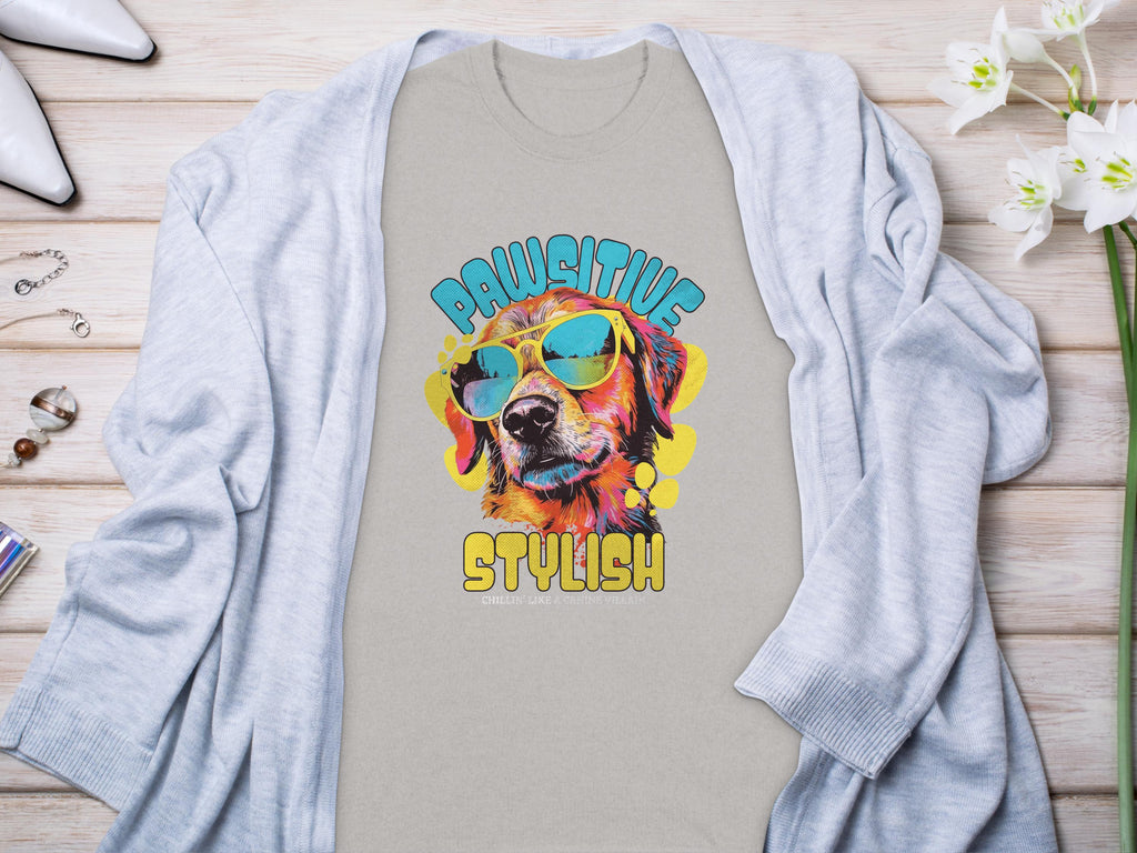 A Garment Graphics gray T-shirt features a vivid dog in sunglasses with Paw-sitive Stylish vibes. The phrase Chillin Like A Canine Villain adds playfulness, complemented by a cardigan, jewelry, and flowers on a wooden surface.