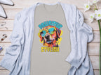 A Garment Graphics gray T-shirt features a vivid dog in sunglasses with Paw-sitive Stylish vibes. The phrase Chillin Like A Canine Villain adds playfulness, complemented by a cardigan, jewelry, and flowers on a wooden surface.