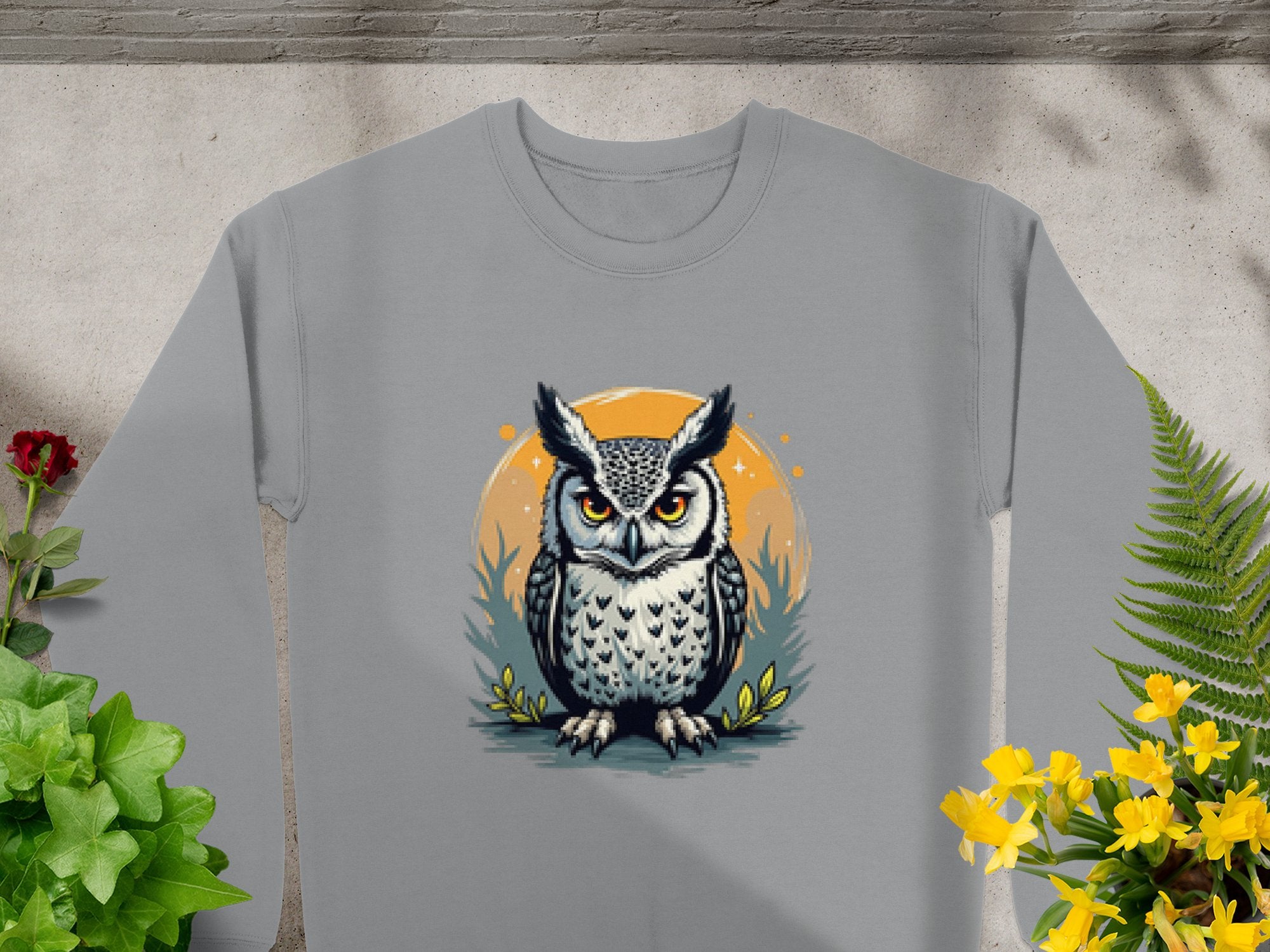 A gray sweatshirt from Garment Graphics showcases an intricate owl design with orange eyes within an orange circle, surrounded by foliage and yellow flowers. Available in unisex sizes for a perfect fit.