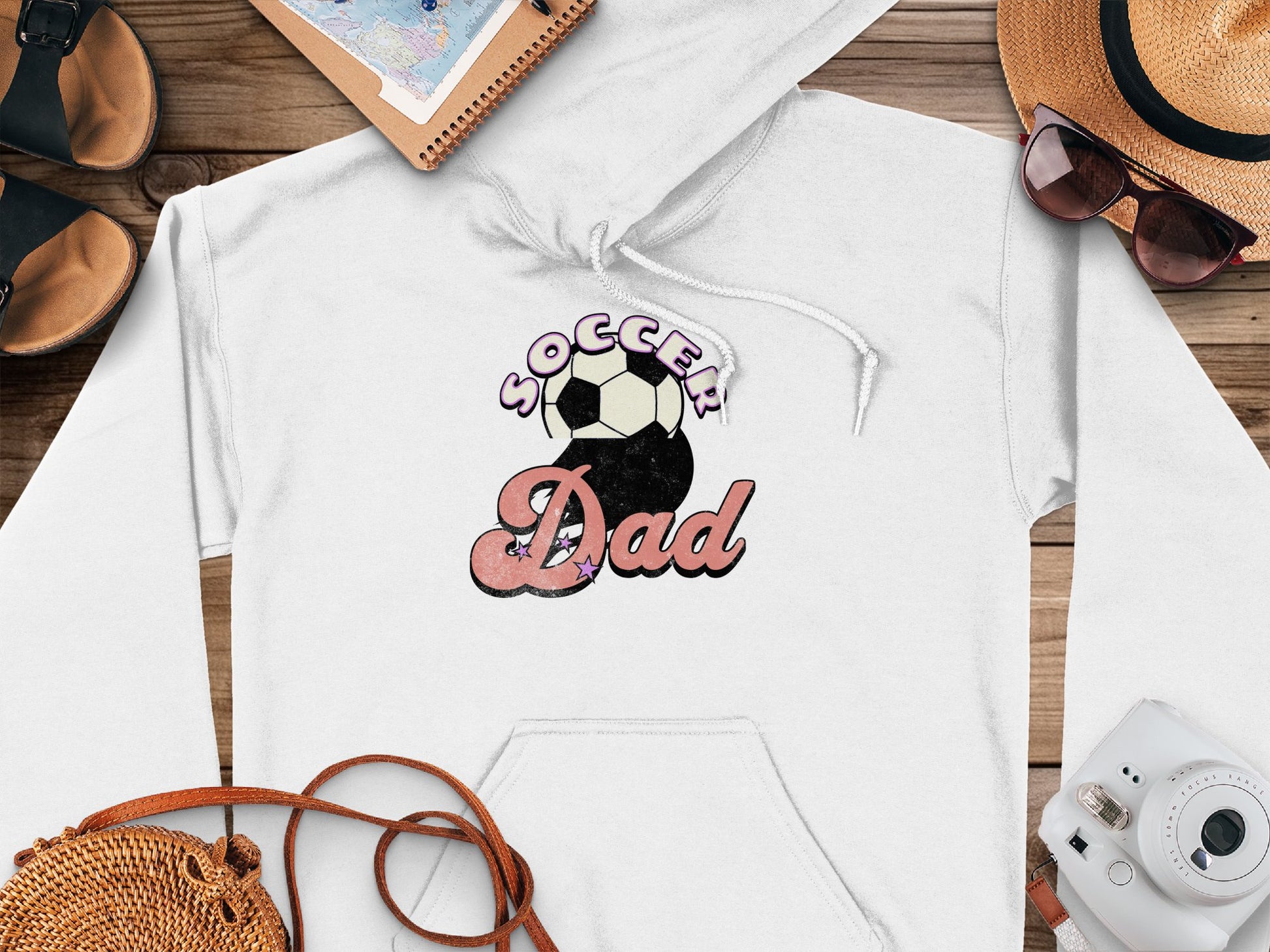 A stylish white hoodie from Garment Graphics features a Soccer Dad print with a soccer ball, displayed on wood beside a hat, sunglasses, notepad, sandals, and camera. Ideal for game days or casual outings.