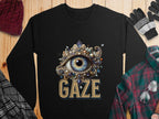 A unisex black graphic sweatshirt from Garment Graphics showcases an ornate eye design embroidered with stones and beads, with GAZE boldly printed in gold underneath. Its displayed on wood surrounded by winter accessories.