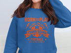 A person wearing a Garment Graphics blue football sweatshirt with orange text and graphics, featuring a soccer ball, lightning bolts, and the words Born to Play, One Goal - One Dream, Young Squad, Football, and Kick. Score. Celebrate! in unisex sizing design.