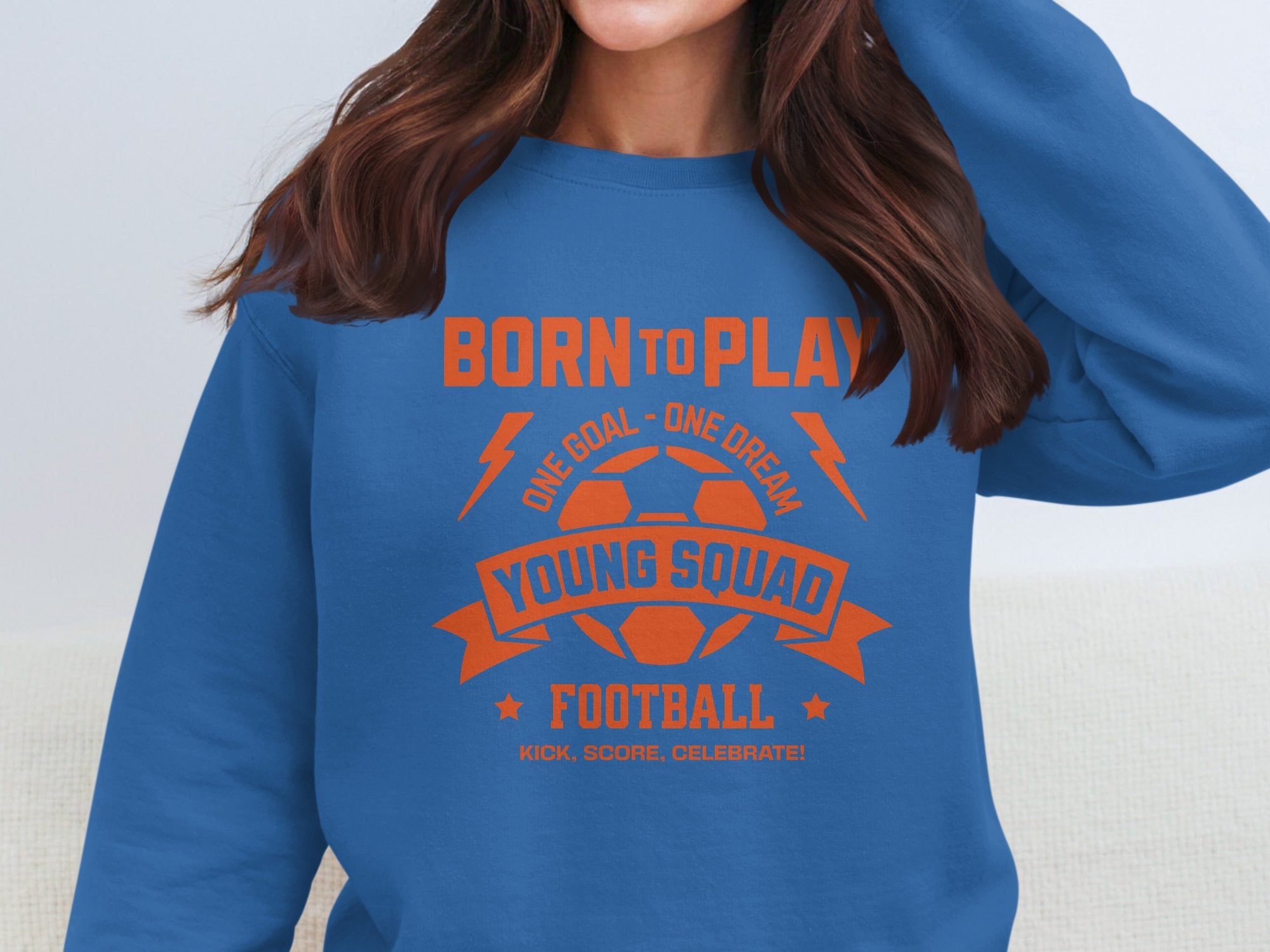 A person wearing a Garment Graphics blue football sweatshirt with orange text and graphics, featuring a soccer ball, lightning bolts, and the words Born to Play, One Goal - One Dream, Young Squad, Football, and Kick. Score. Celebrate! in unisex sizing design.