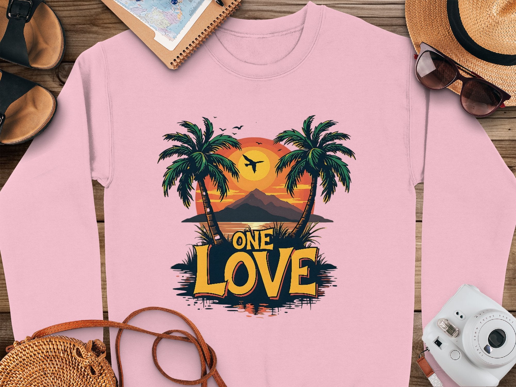 A pink Garment Graphics sweatshirt featuring a One Love Sunset beach scene with palm trees and a plane at dusk lies on wood, accompanied by travel essentials: hat, camera, sunglasses, shoes, and notebook—ideal for relaxed adventures.