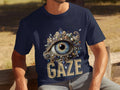 A person wears a navy blue Garment Graphics t-shirt featuring a jeweled ornate eye illustration with GAZE printed below. This classic fit tee, adorned with unique designs, sits perfectly on them as they relax on a wooden bench outdoors.