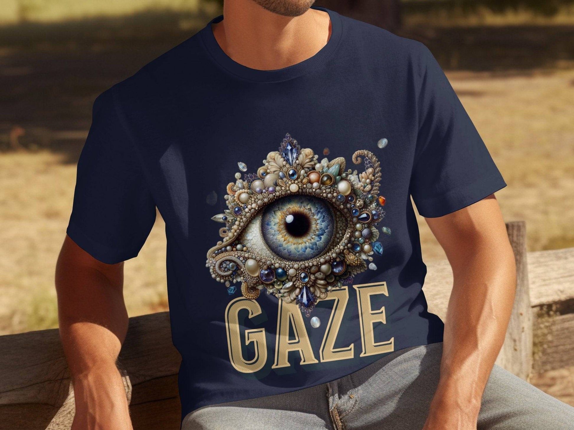 A person wears a navy blue Garment Graphics t-shirt featuring a jeweled ornate eye illustration with GAZE printed below. This classic fit tee, adorned with unique designs, sits perfectly on them as they relax on a wooden bench outdoors.