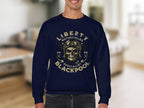 A person wearing a navy Garment Graphics sweatshirt with a skull in a vintage pilot helmet graphic, biker-style. Text says Liberty Motorcycle Specialist Blackpool 2023 with two crossed wrenches, against a softly focused room backdrop.