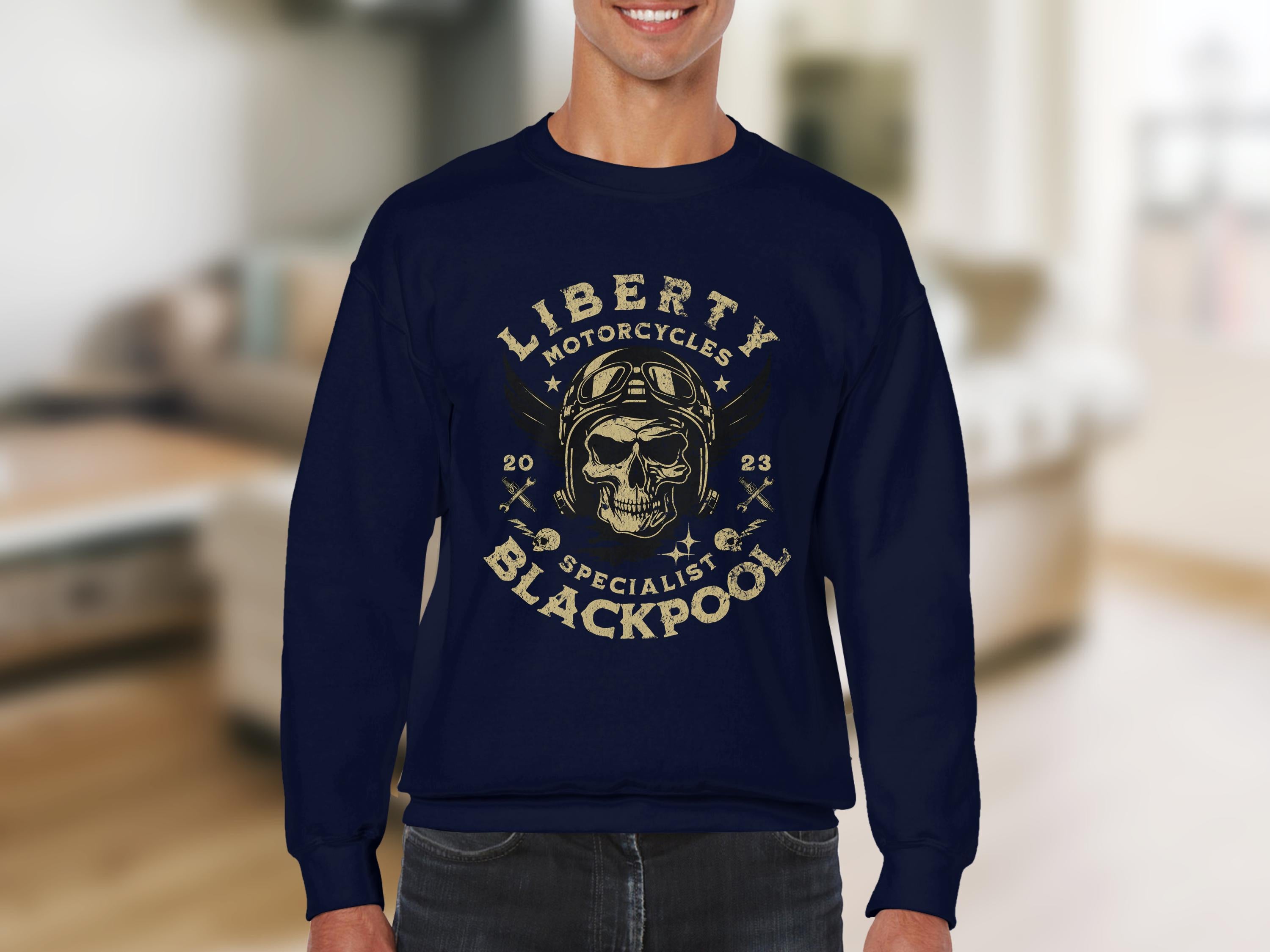 A person wearing a navy Garment Graphics sweatshirt with a skull in a vintage pilot helmet graphic, biker-style. Text says Liberty Motorcycle Specialist Blackpool 2023 with two crossed wrenches, against a softly focused room backdrop.
