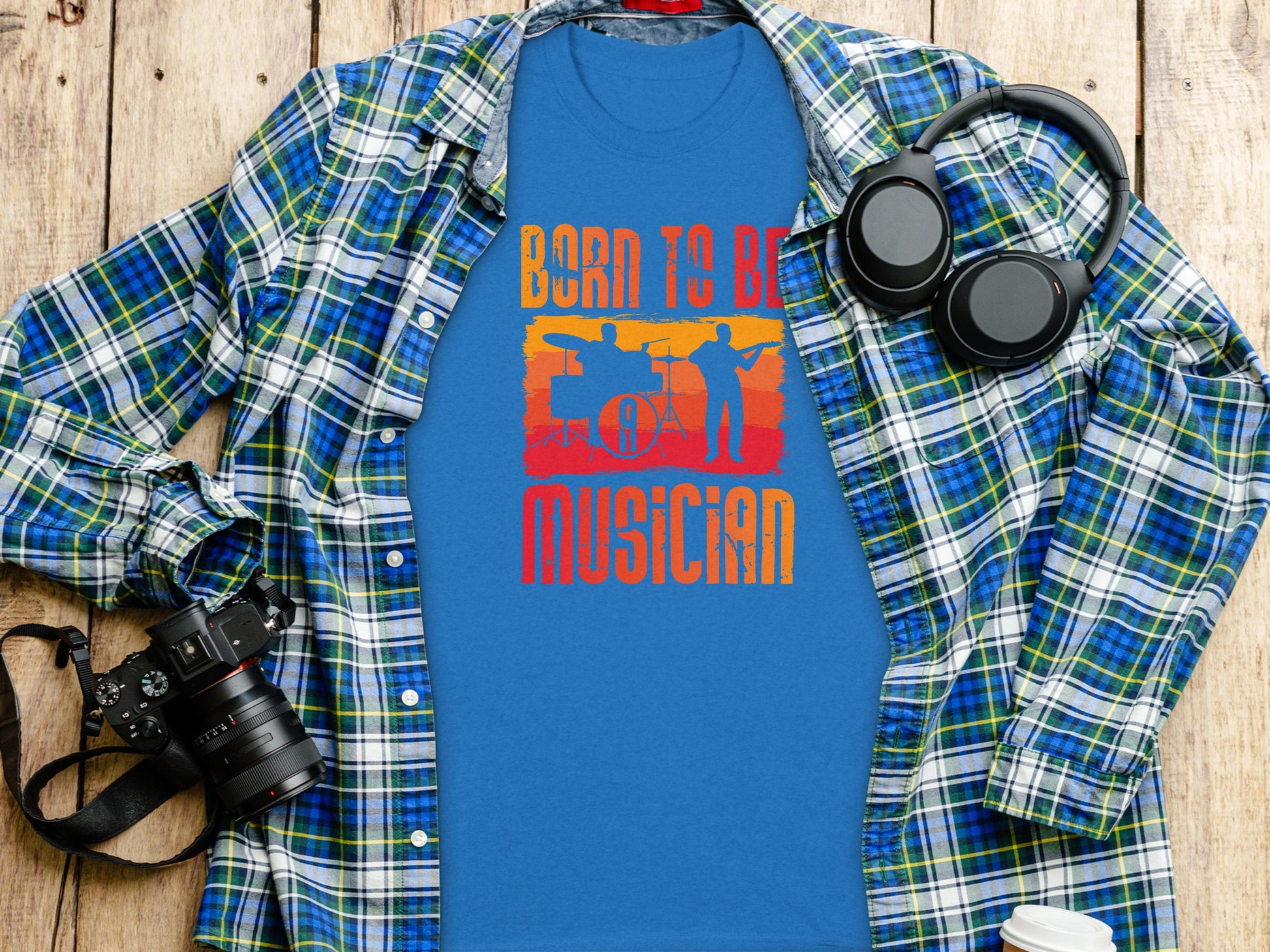 A blue Born to Be a Musician T-shirt by Garment Graphics, showcasing a drummer silhouette, is displayed flat. Ideal for music enthusiasts, this tee complements a blue and green plaid shirt, headphones, camera, and coffee cup on the wooden surface.