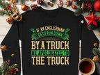 A unisex black sweatshirt from Garment Graphics is on a wooden surface with festive Christmas decor, featuring the humorous text, If an Englishman gets run down by a truck he apologizes to the truck in bold green and beige lettering.