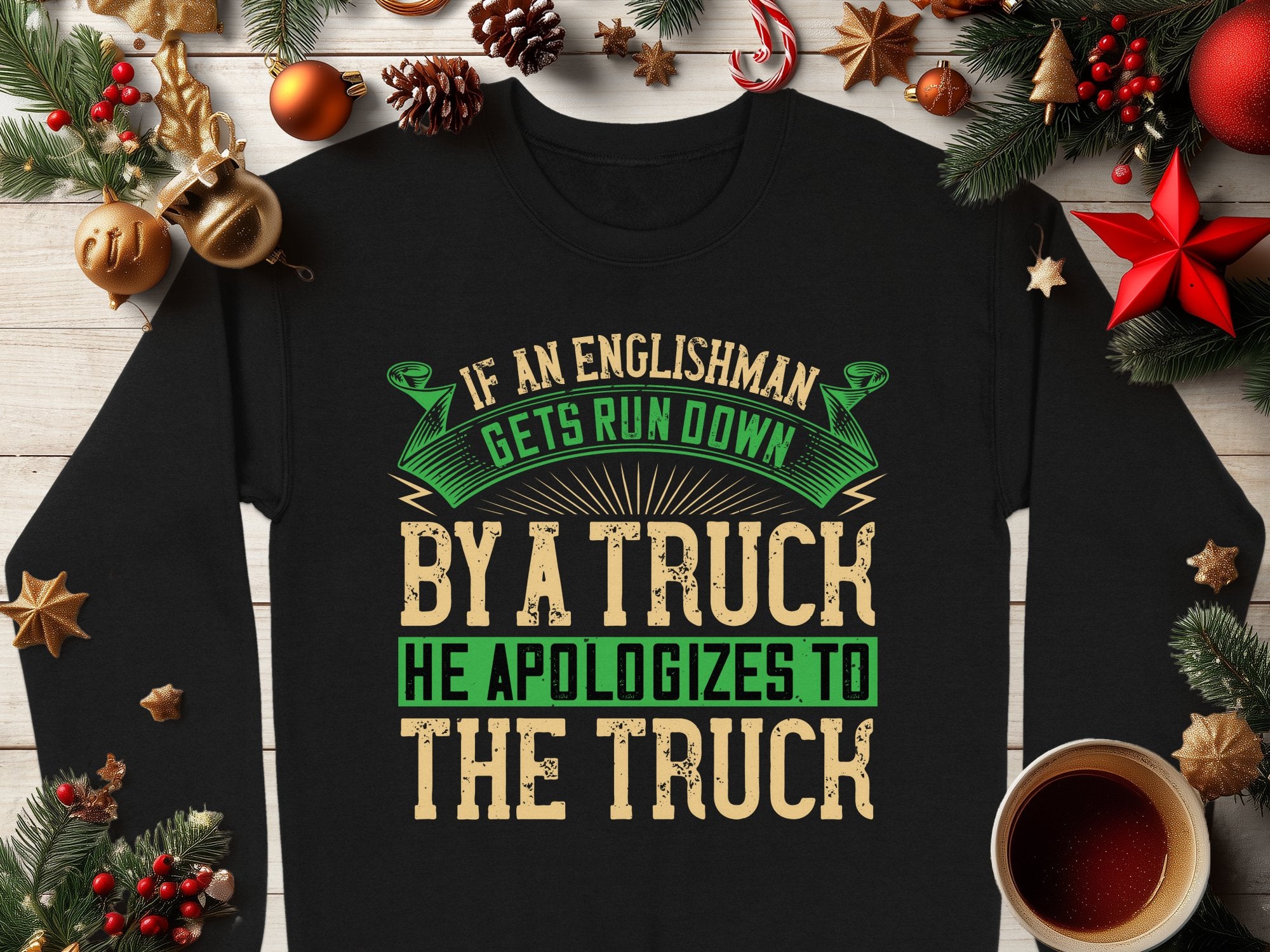 A unisex black sweatshirt from Garment Graphics is on a wooden surface with festive Christmas decor, featuring the humorous text, If an Englishman gets run down by a truck he apologizes to the truck in bold green and beige lettering.