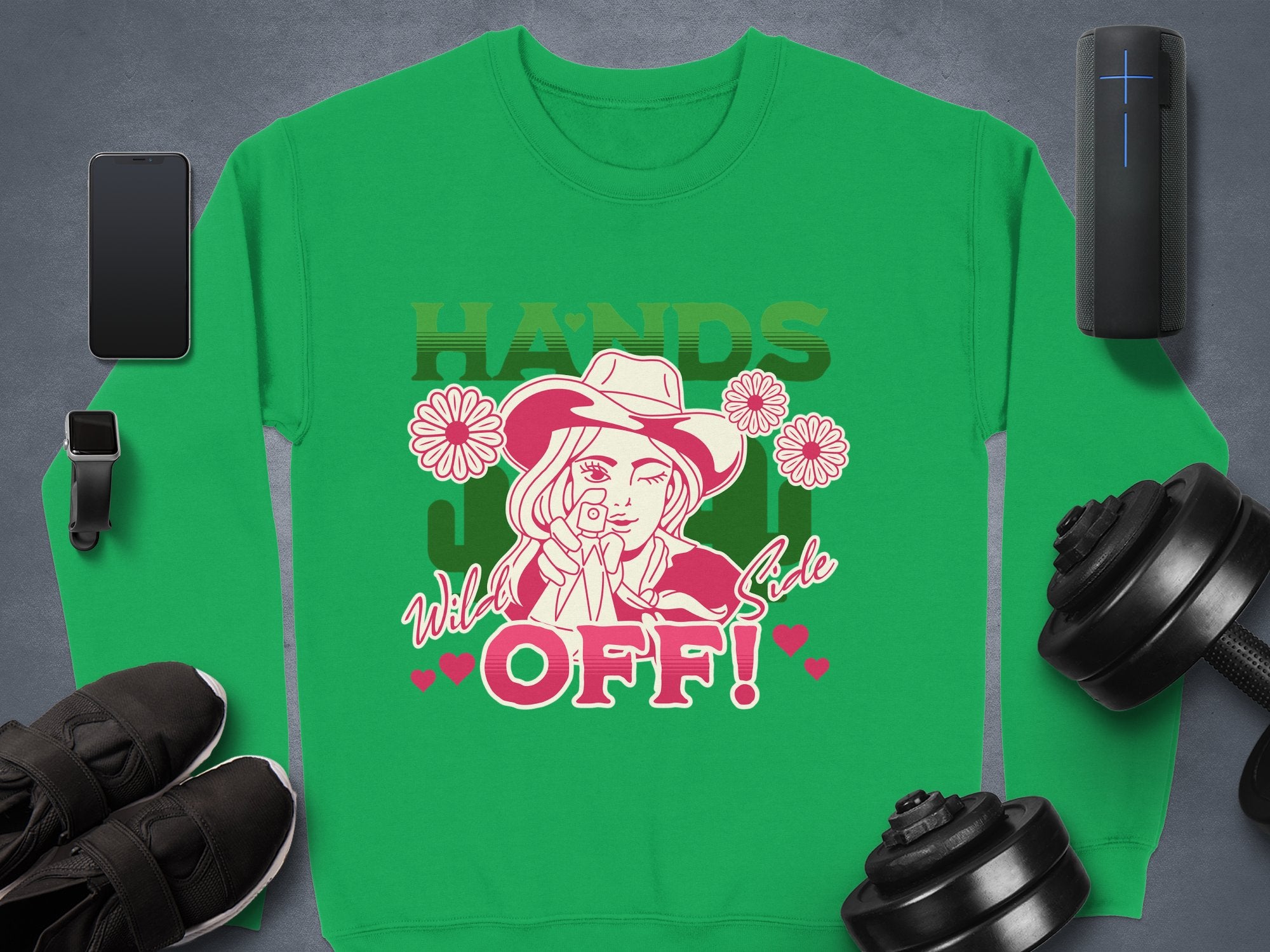 Green loose-fit sweatshirt by Garment Graphics, featuring a line drawing of a person in a hat surrounded by flowers with the texts HANDS OFF! and Wild West Style. Displayed flat alongside essentials like a phone, shoes, dumbbells, charging cable, and speaker.