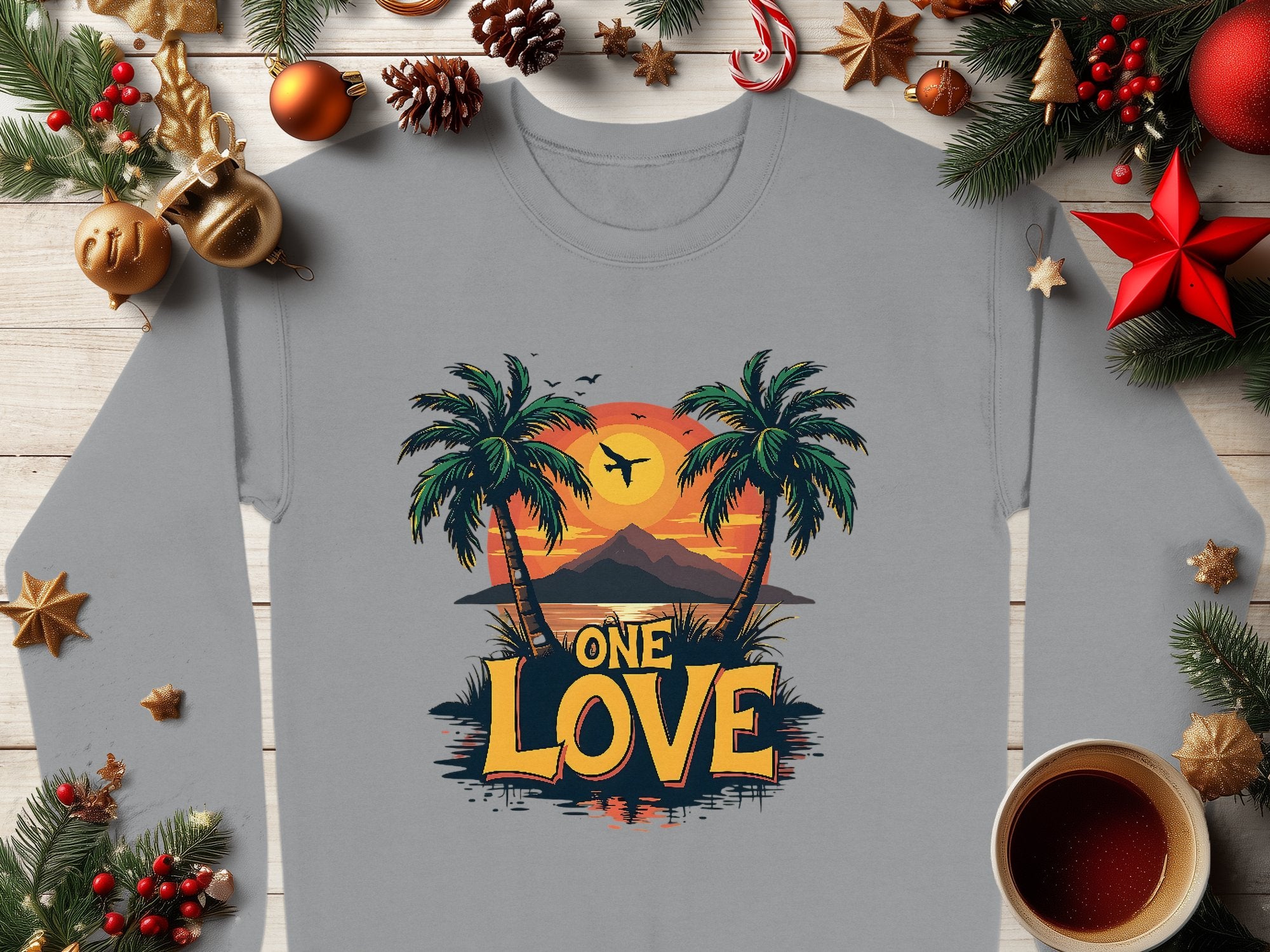 The Garment Graphics One Love Sweatshirt, in unisex sizing, features a gray long-sleeve design with a sunset between palm trees, an airplane, mountains, and festive holiday elements like pine branches, ornaments, and a candle.
