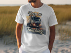 A truck enthusiast wears a Garment Graphics white t-shirt with a large truck graphic on a road, Keep on Trucking text, and a grassy field under a clear sky backdrop.