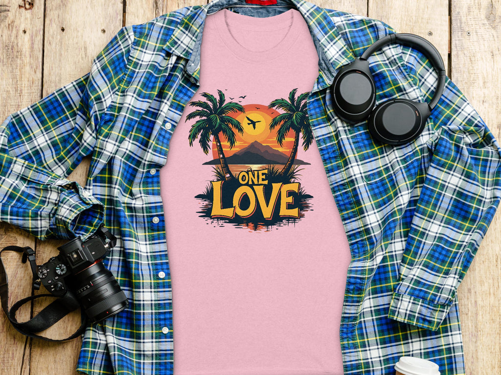A Garment Graphics pink T-shirt featuring a One Love tropical sunset design with palm trees and mountains lies on a wooden surface, surrounded by a blue-green plaid shirt, headphones, camera, and takeaway coffee cup—perfect for beach enthusiasts.