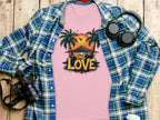 A Garment Graphics pink T-shirt featuring a One Love tropical sunset design with palm trees and mountains lies on a wooden surface, surrounded by a blue-green plaid shirt, headphones, camera, and takeaway coffee cup—perfect for beach enthusiasts.