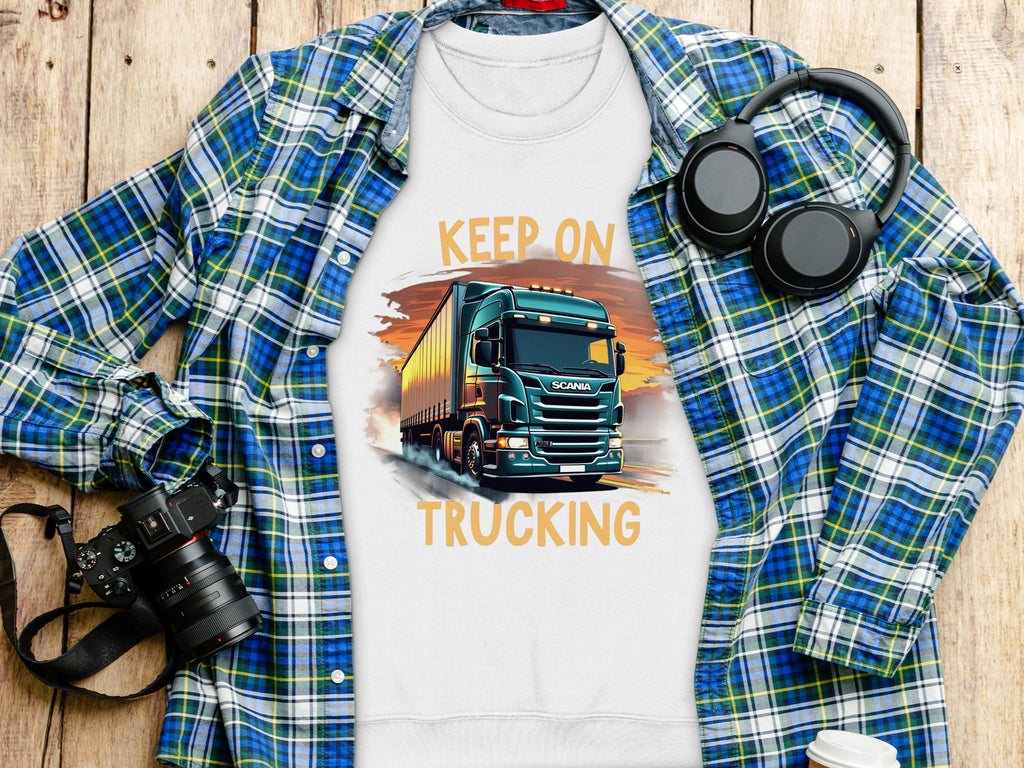 A Garment Graphics white t-shirt with a bold truck design and Keep On Trucking text is displayed on a wooden surface, alongside a blue plaid shirt, black headphones, a black camera, and a white lidded cup.