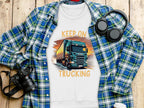 A Garment Graphics white t-shirt with a bold truck design and Keep On Trucking text is displayed on a wooden surface, alongside a blue plaid shirt, black headphones, a black camera, and a white lidded cup.