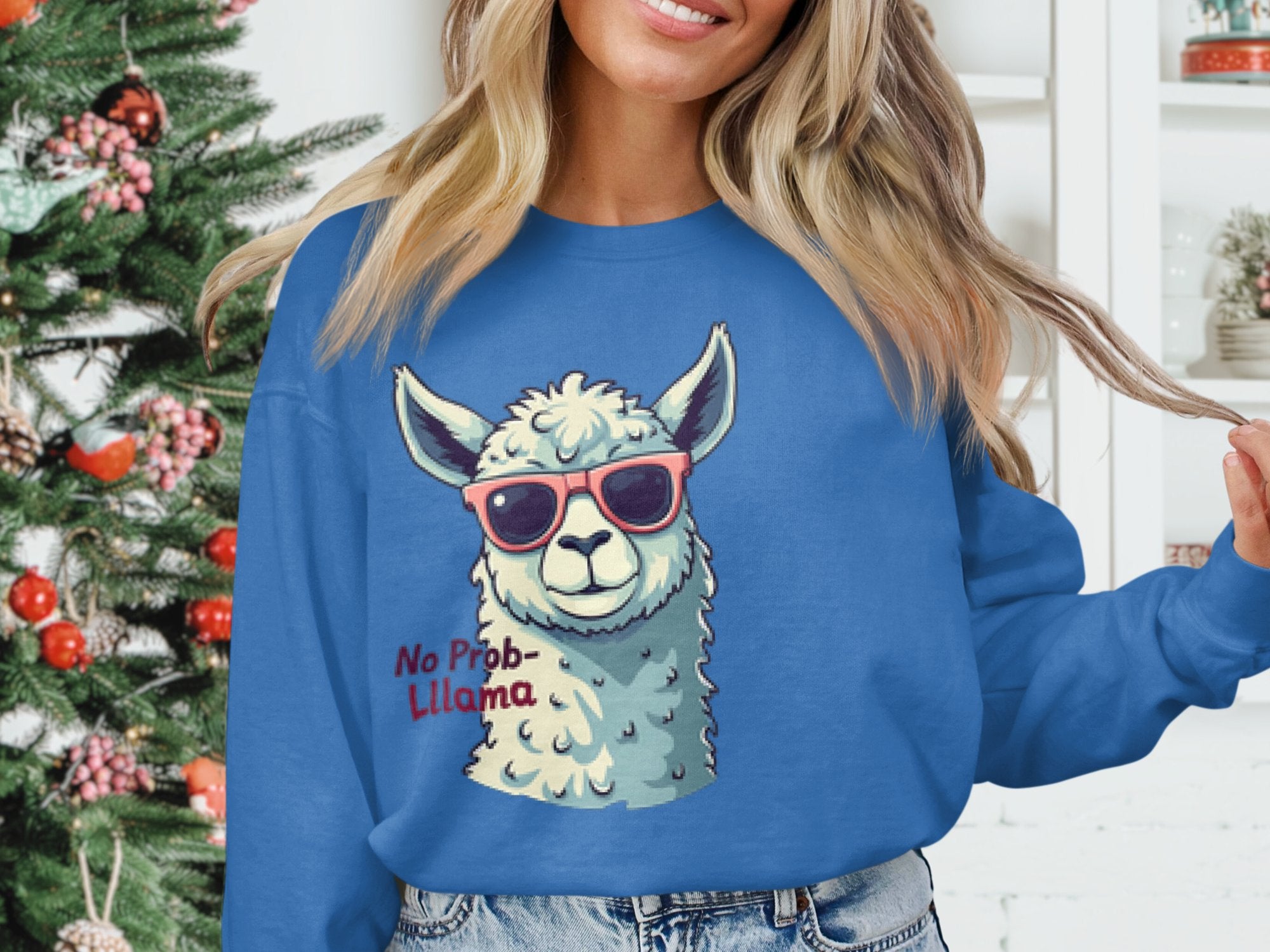 Near a Christmas tree adorned with red and white ornaments, someone wears Garment Graphics humorous No Prob-Llama sweatshirt, featuring a llama with sunglasses. The unisex sizing makes it an ideal festive choice for anyone.